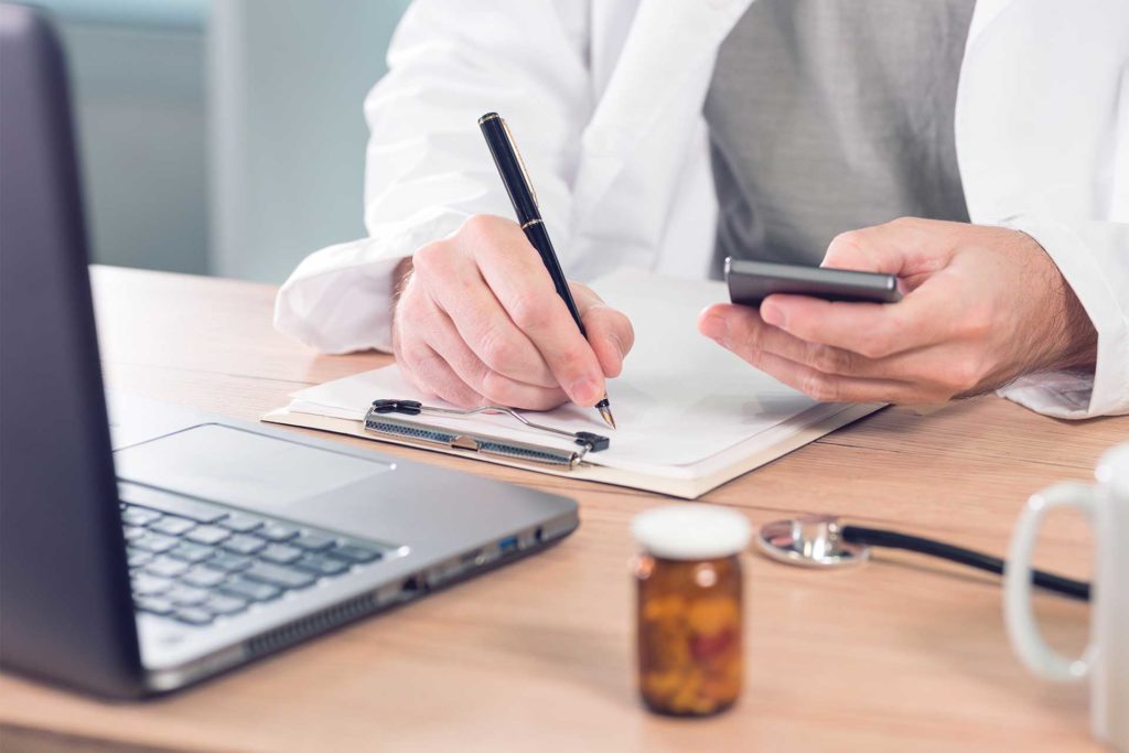 doctor writing on clipboard in front of laptop