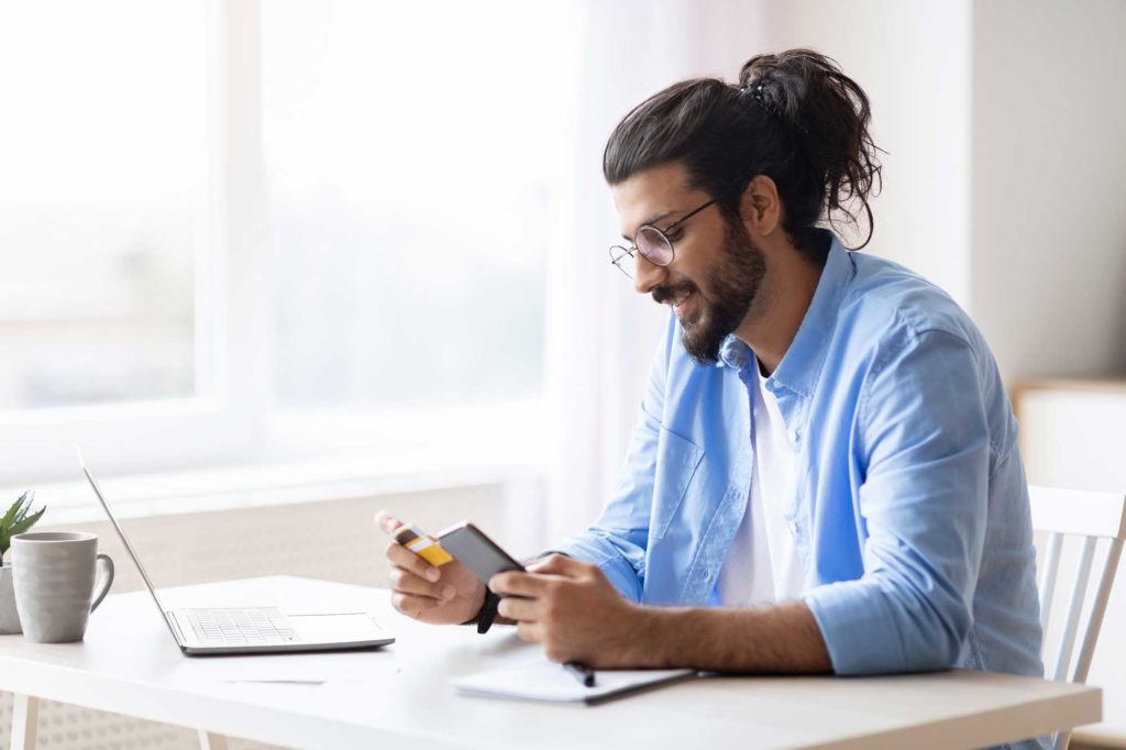 young-arab-guy-using-smartphone-and-credit-card-at-computer