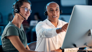 Two BPO customer service representatives wearing headsets working together at a computer in a modern office setting