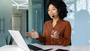 Smiling customer service representative wearing a headset and speaking on a video call from a modern office, using a laptop to assist clients